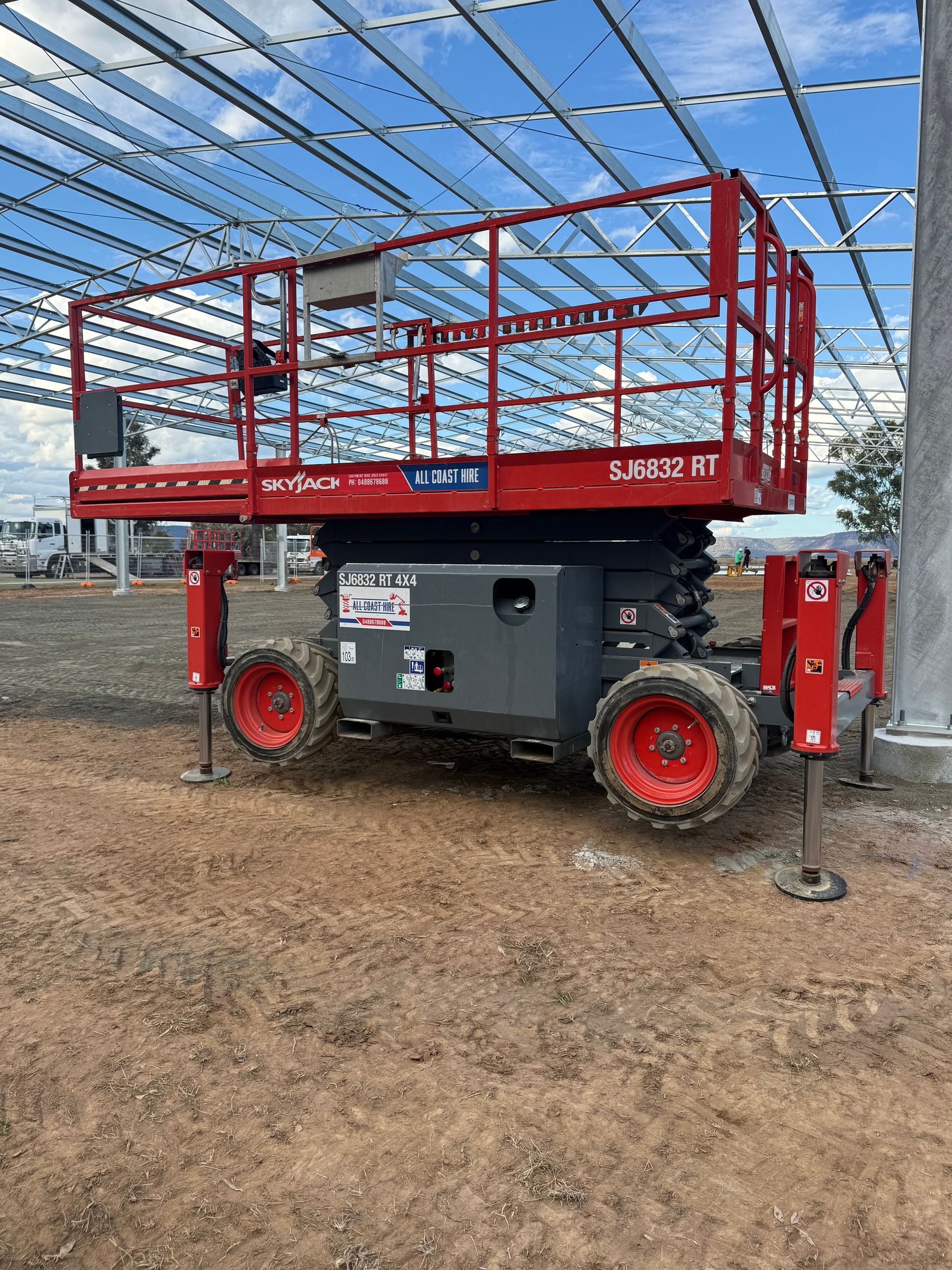 A red rough-terrain scissor lift with extended stabilizer legs sits on a dirt construction site under metal framework.