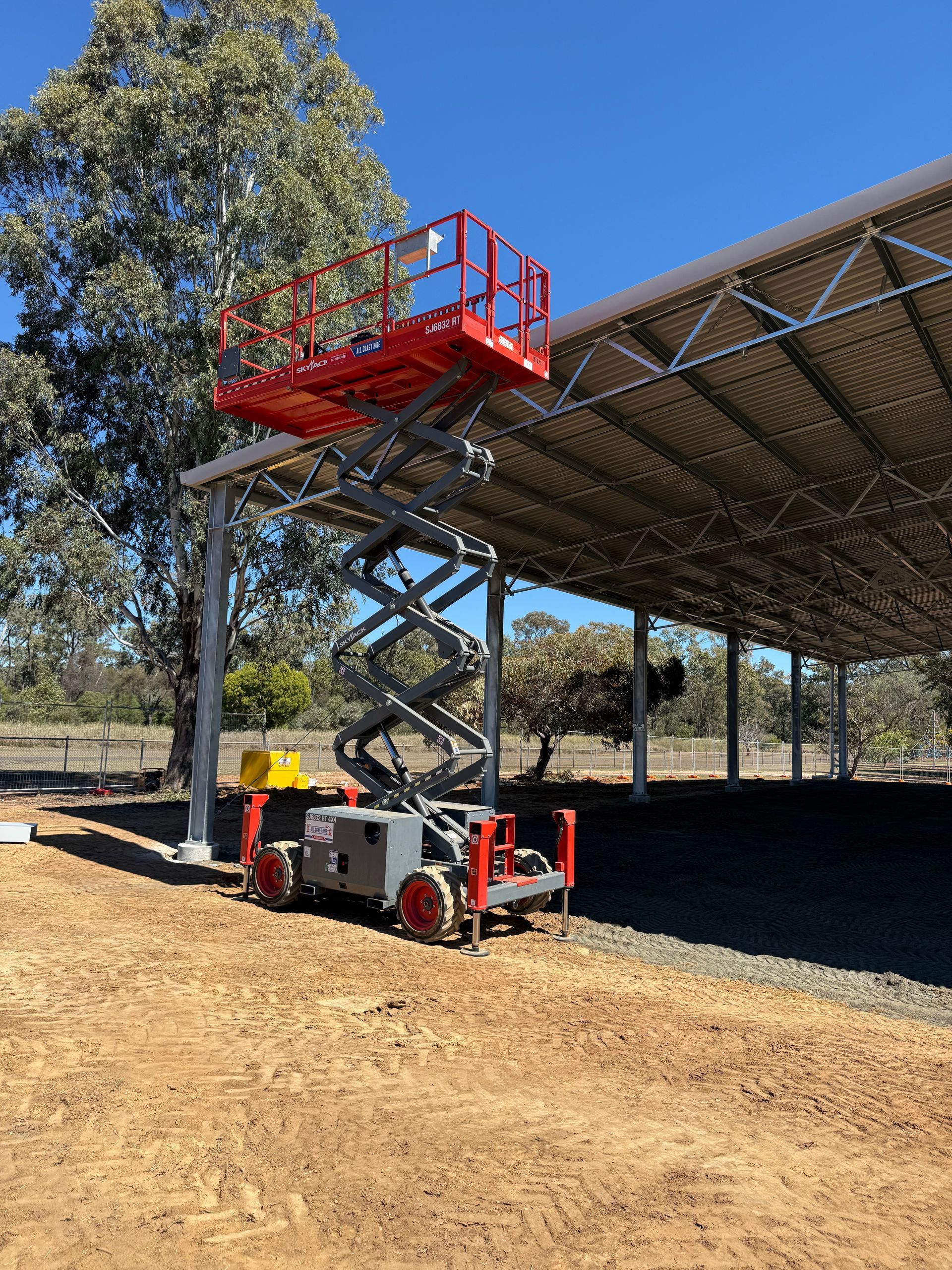 A red scissor lift is extended to its maximum height next to a large, open-air metal structure on a sunny day.