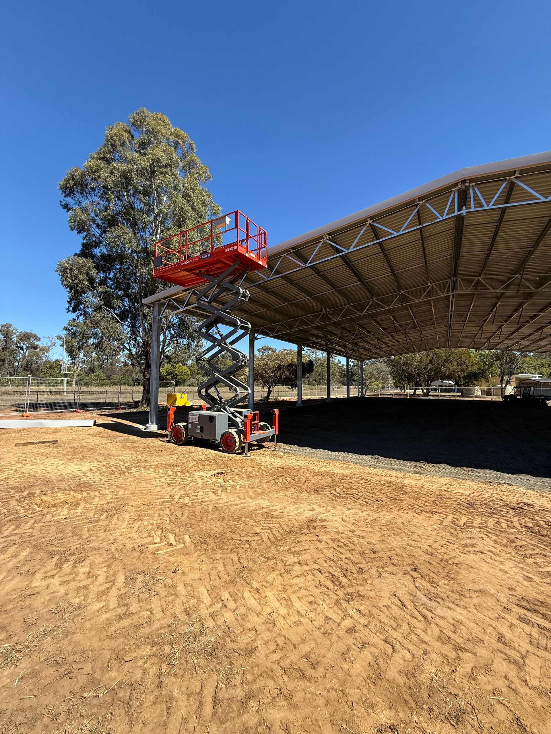 A red scissor lift is extended next to a large, open-air metal roof structure on a dirt lot under a clear blue sky.