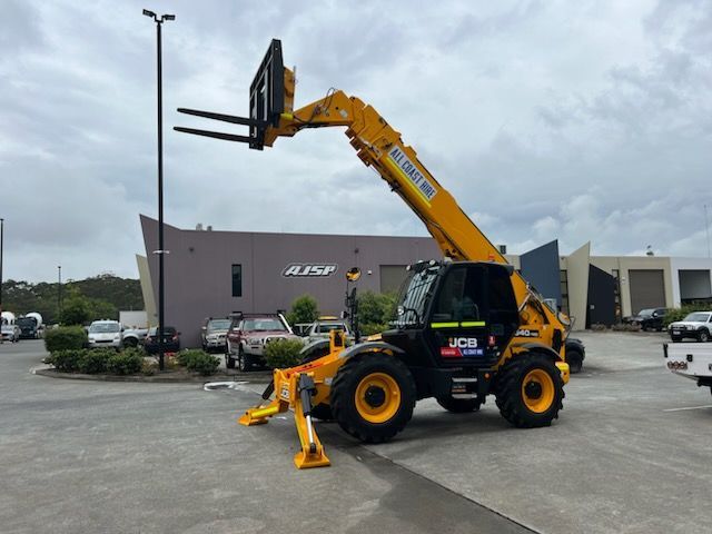 A yellow JCB telescopic handler with extended forks and deployed outriggers parked in a parking lot.