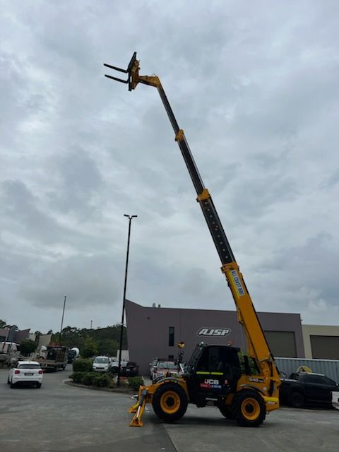 A yellow telescopic handler with an extended boom and forks, parked in an outdoor industrial lot under a cloudy sky.