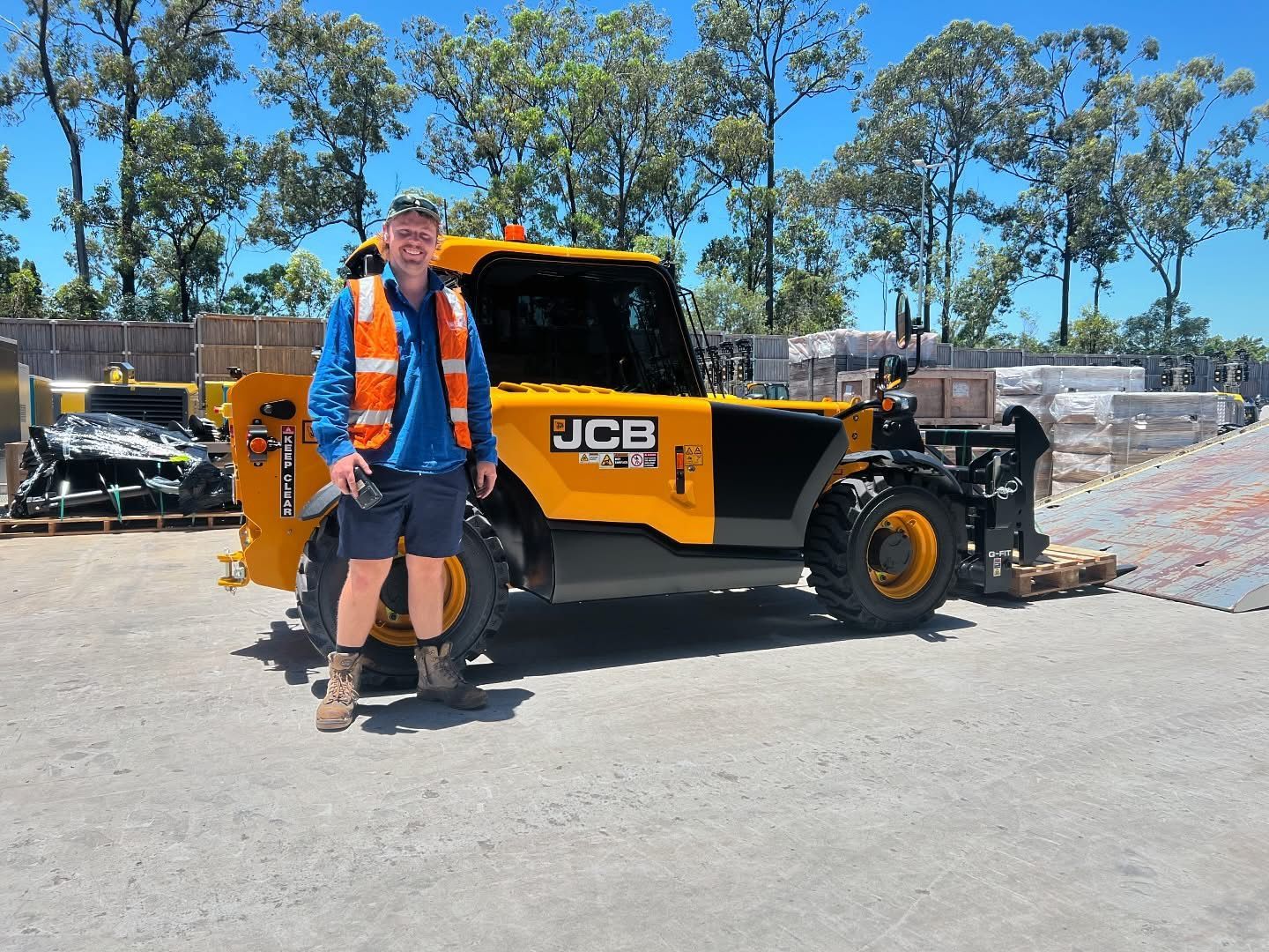 A person in a blue shirt and orange high-visibility vest standing in front of a yellow JCB telescopic handler outdoors.