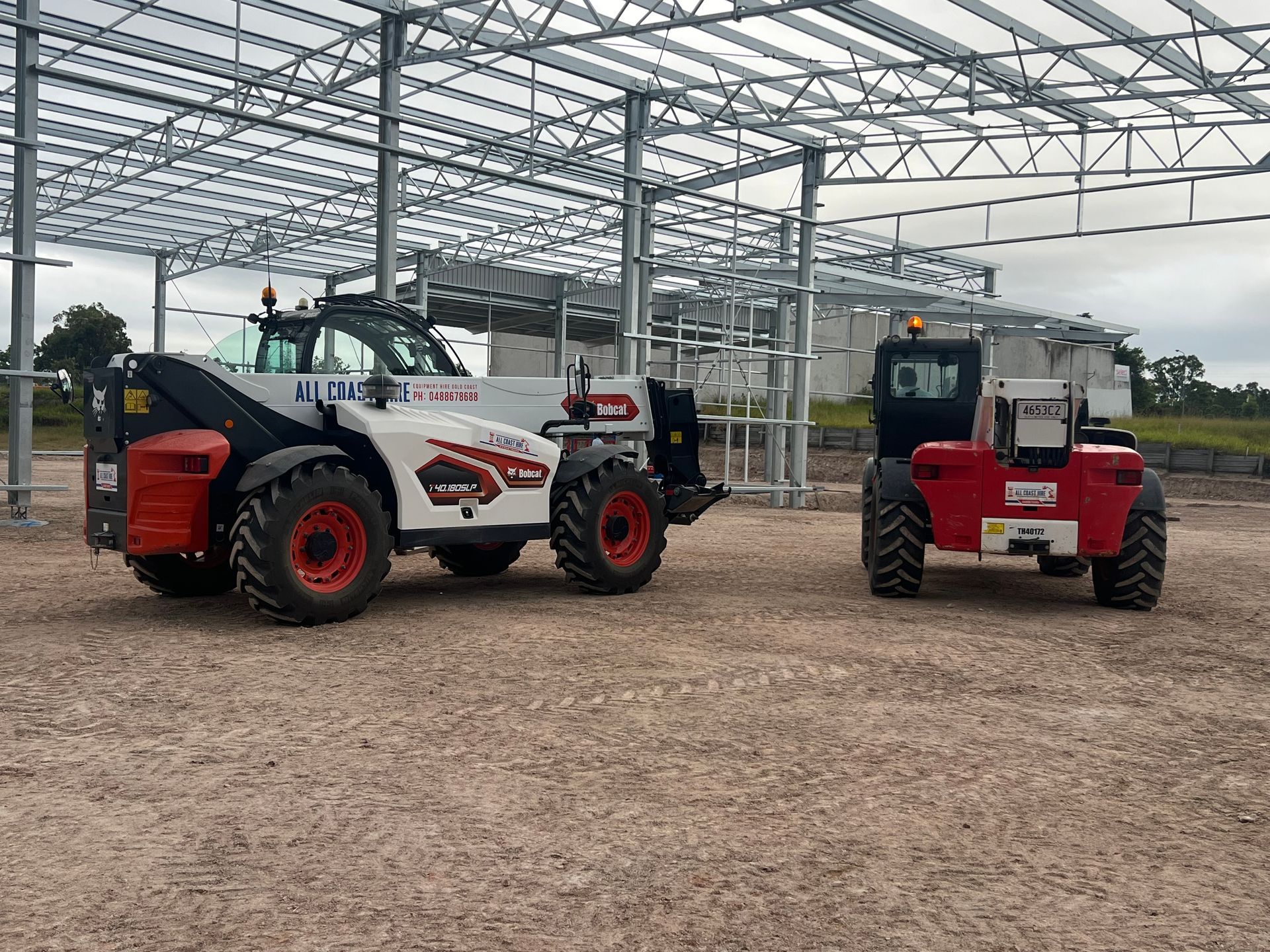 Two Dieci telehandlers, one white and one red, parked on a dirt lot in front of a steel building framework.
