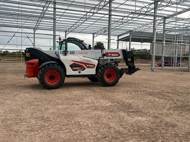 A white and red Bobcat telehandler sits on a dirt lot in front of a steel building frame under construction.