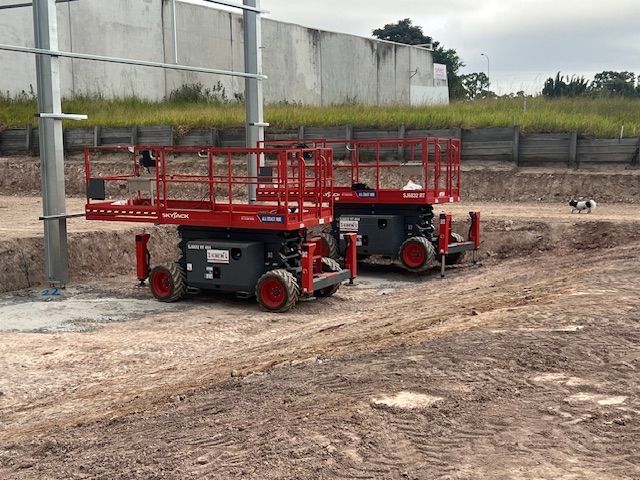 Two red scissor lifts parked on a dirt construction site next to a metal pole, with a concrete wall in the background.
