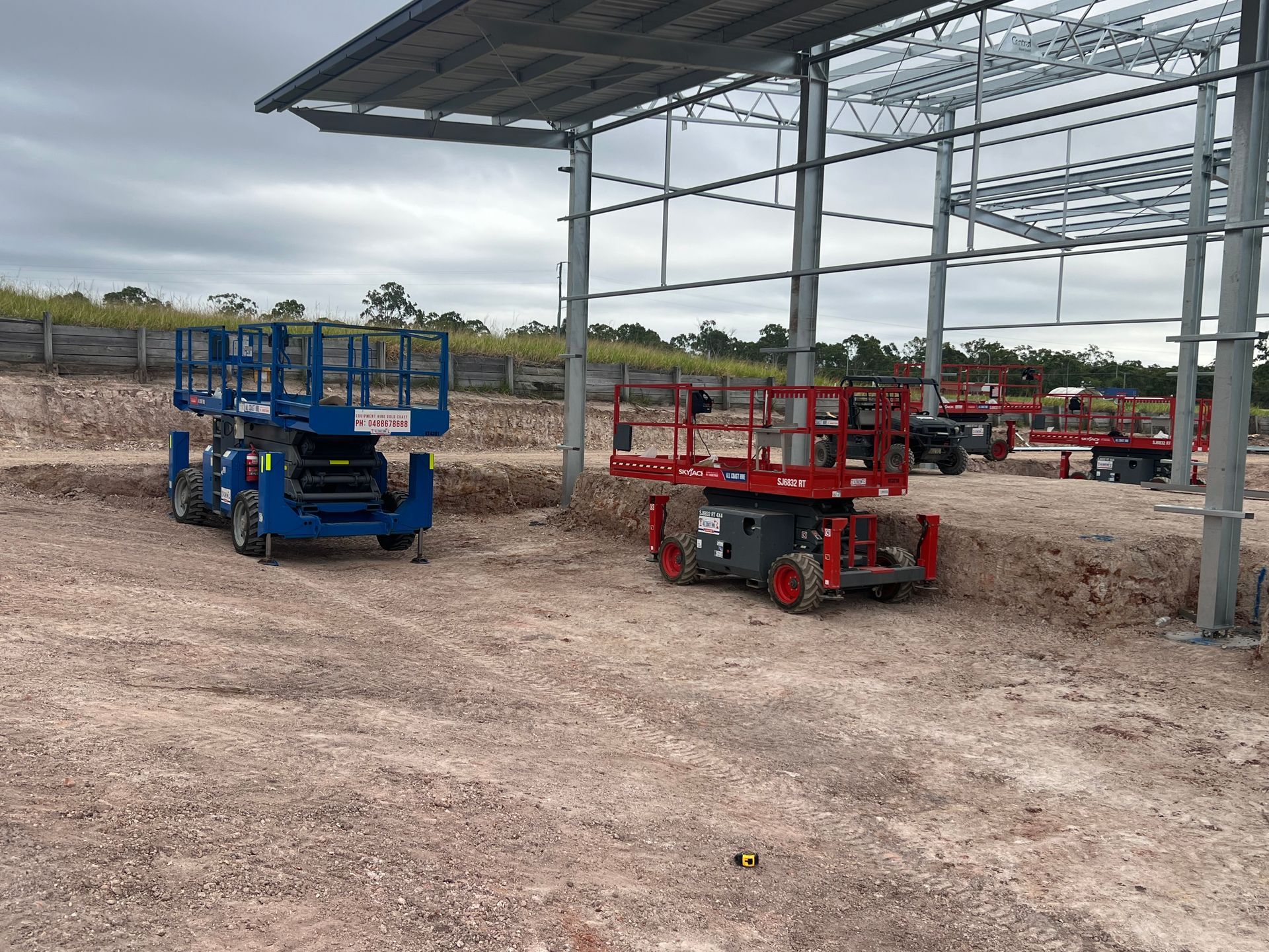 Two scissor lifts, one blue and one red, parked on a gravel construction site beneath a metal structure.