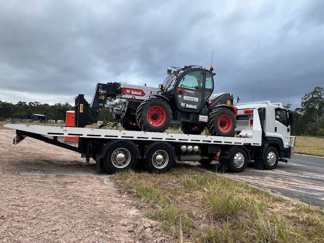 A white flatbed tow truck transporting a red and black Bobcat telehandler on a gravel road under a cloudy sky.