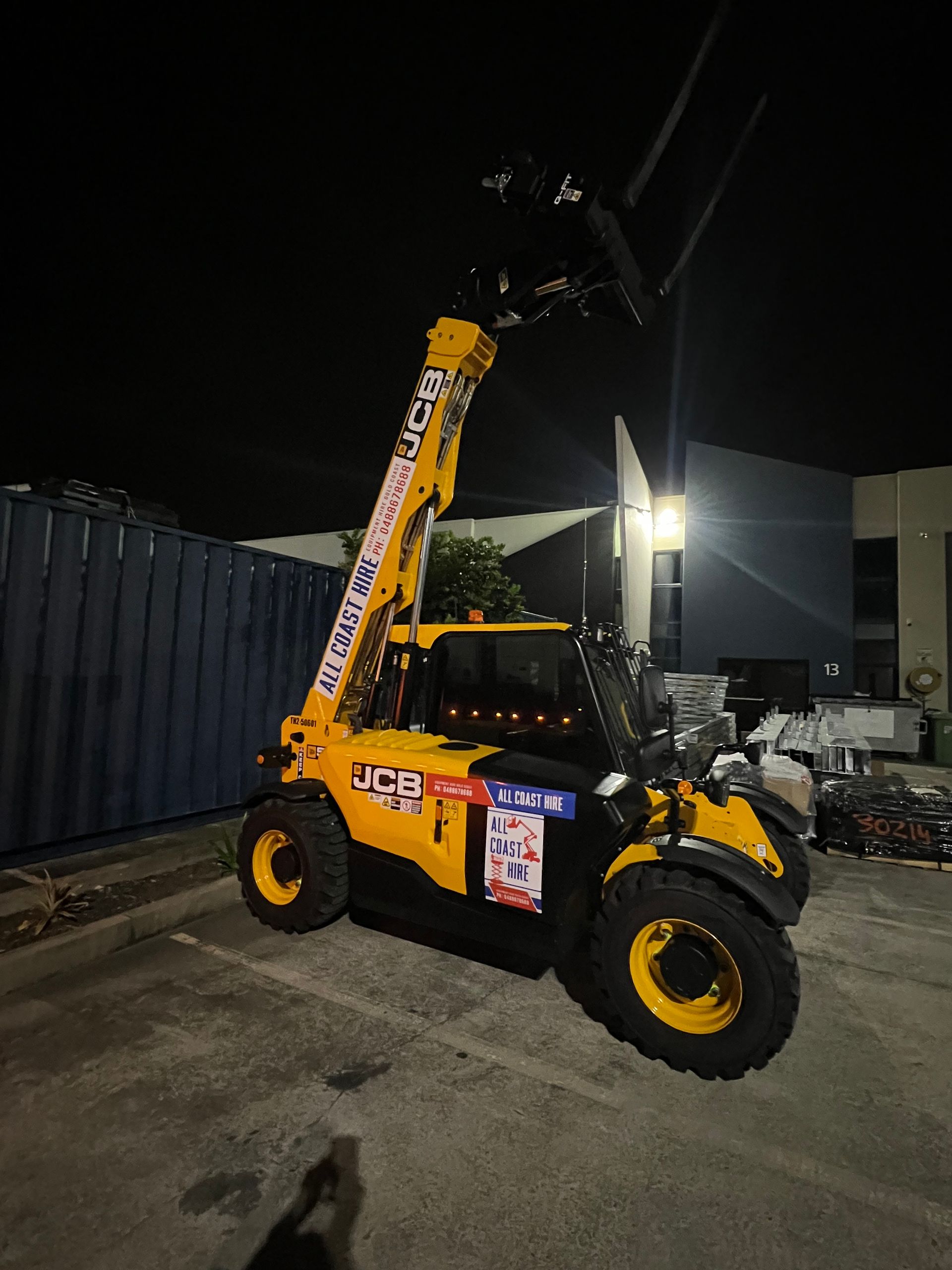 A yellow JCB telehandler parked outdoors at night with its boom extended upward.