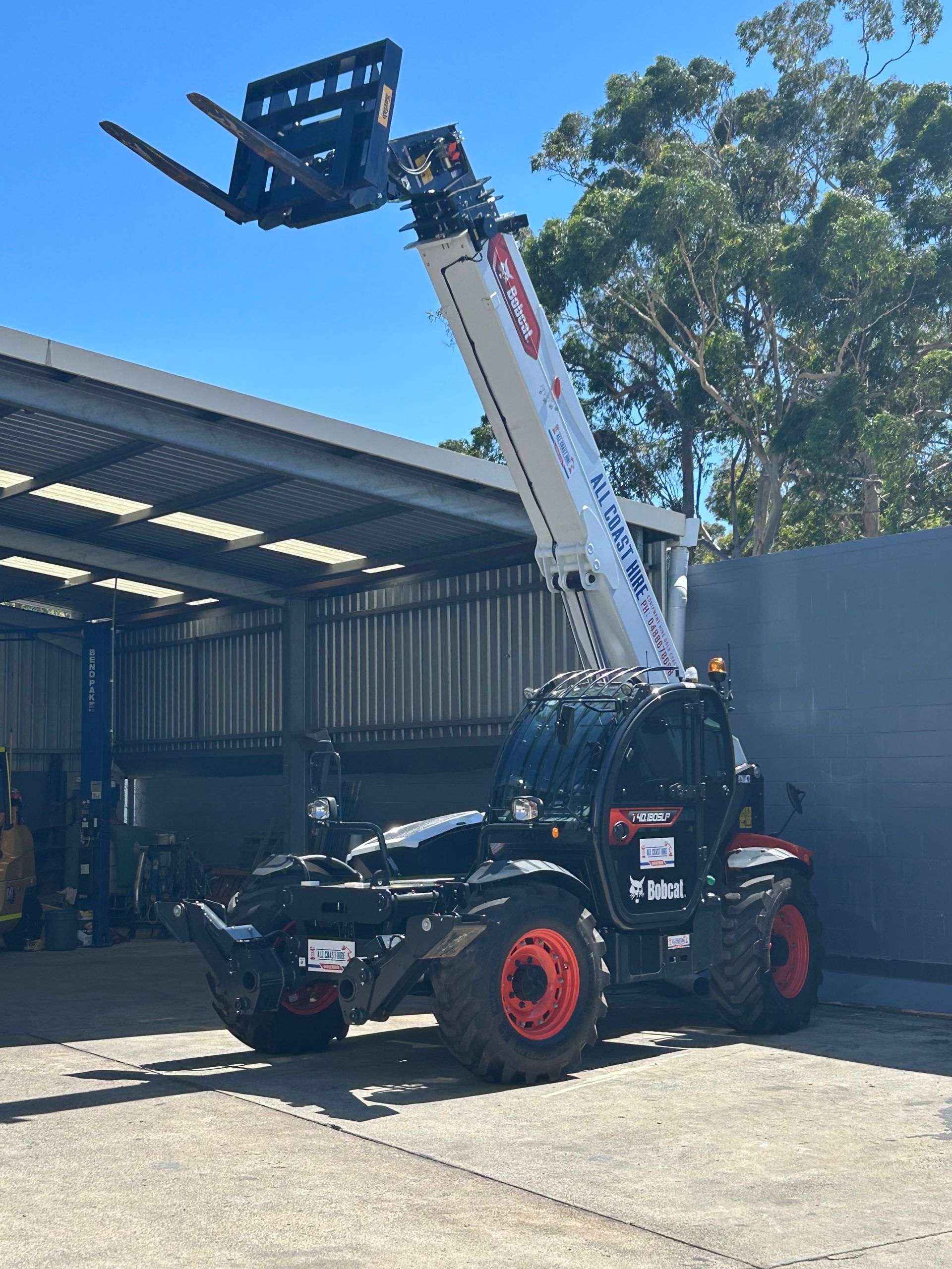 A white and black telescopic forklift with extended forks parked on a concrete surface under a shed roof.