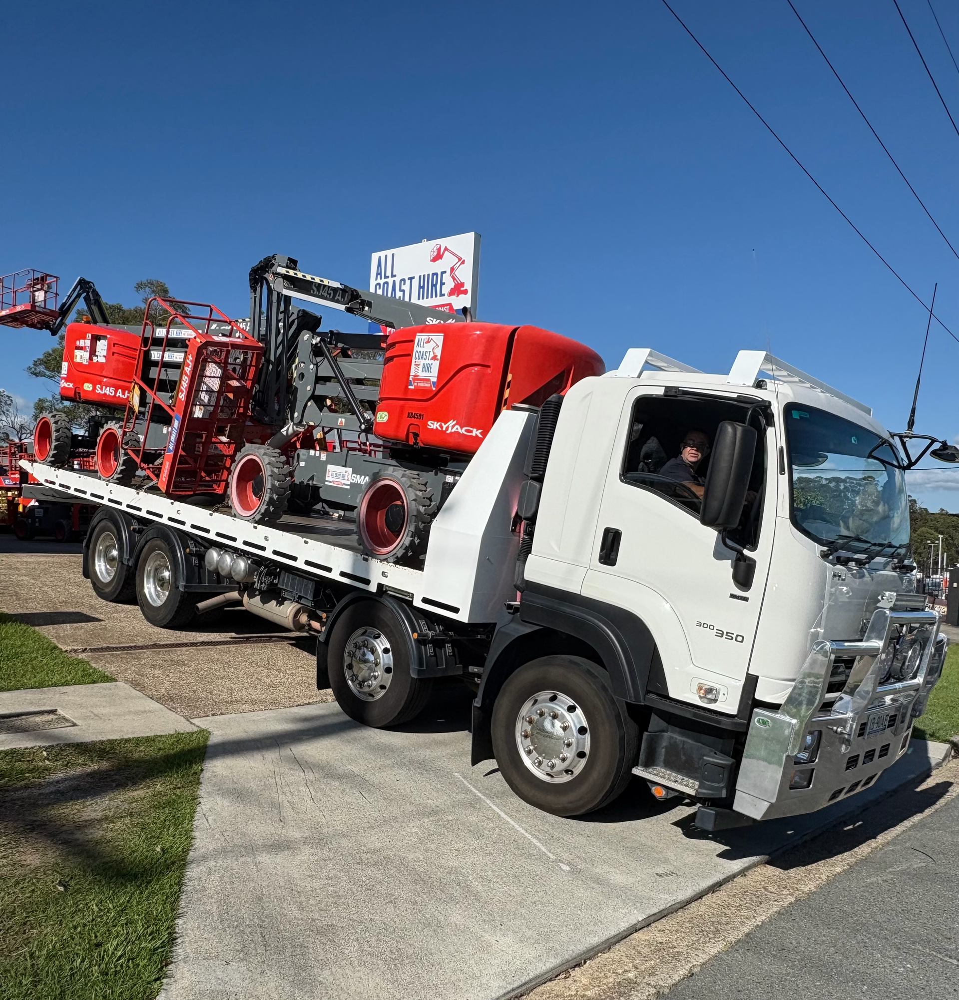 A white flatbed tow truck transporting two red construction boom lifts on a sunny day.