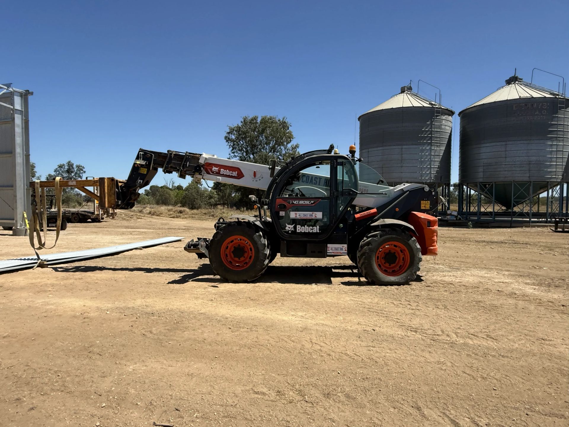Black-and-red telehandler with extended boom on a dirt site near a silo and excavator