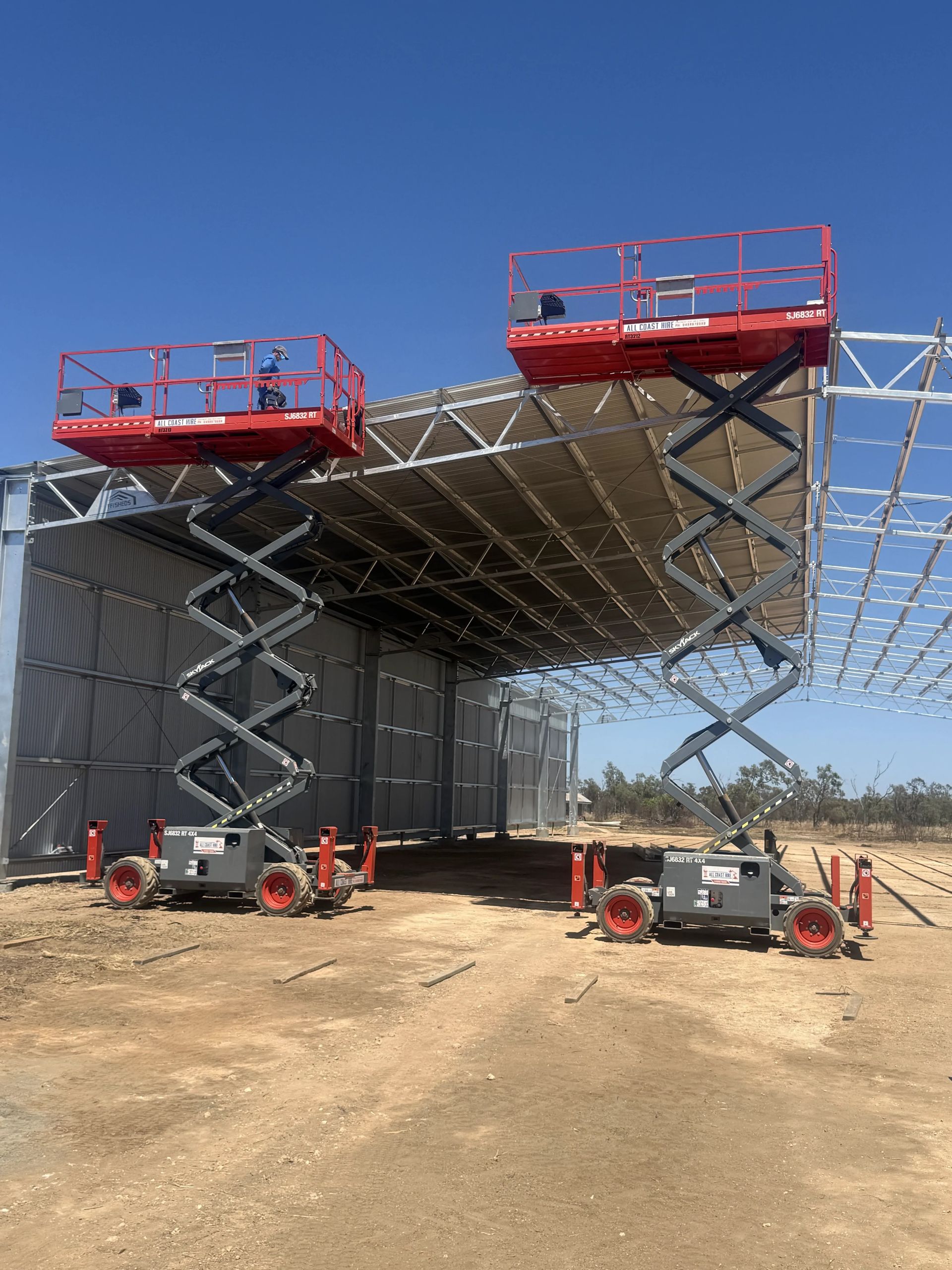 Telehandler lifting a pipe in a farmyard beside grain silos under a clear blue sky