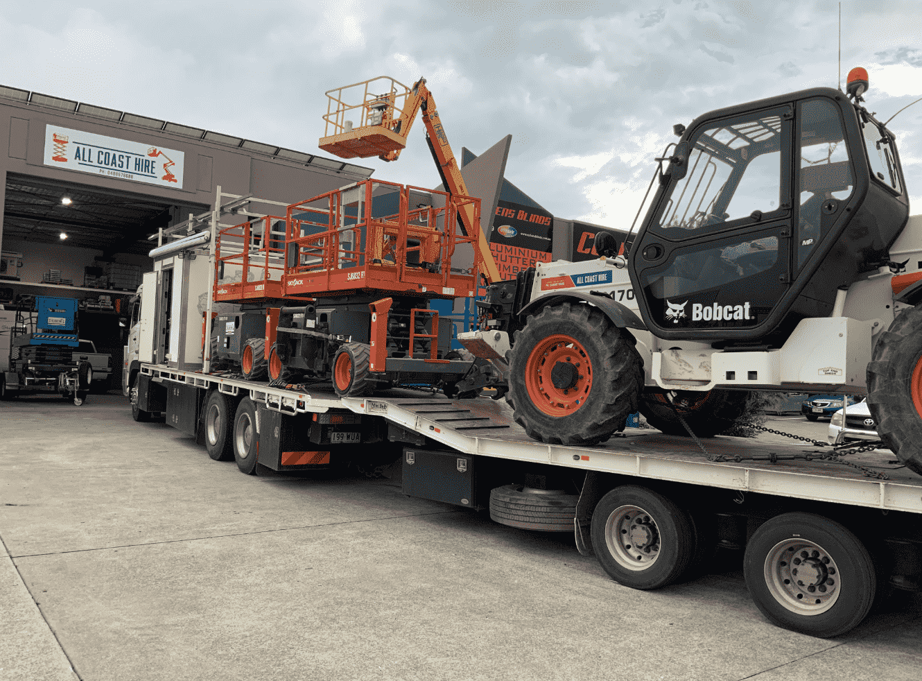 A flatbed truck transporting an orange aerial lift and a white Bobcat telehandler outside a warehouse.