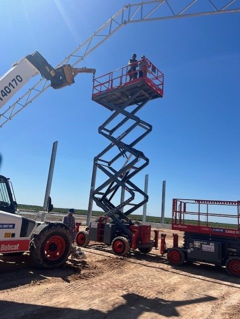 Two people stand on an elevated scissor lift on a dirt construction site near a Bobcat telehandler under a blue sky.