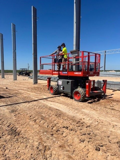 Two people stand in a red scissor lift on a dirt construction site next to tall concrete pillars under a blue sky.
