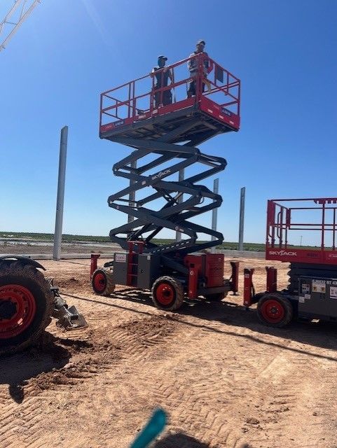 Two workers standing in an elevated red scissor lift on a dirt construction site under a clear blue sky.
