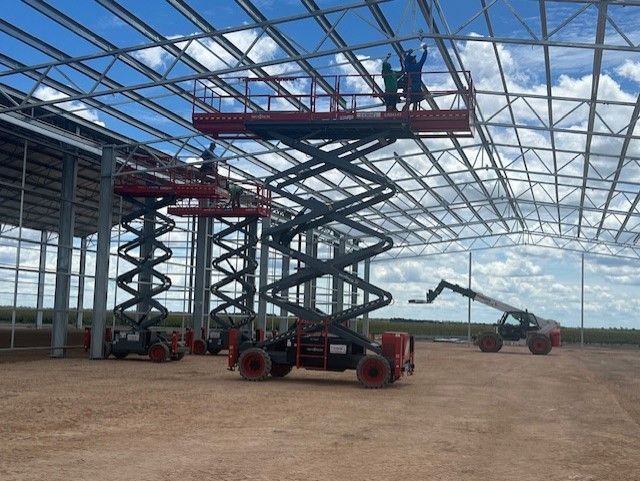 Workers on red scissor lifts install metal roof beams in a large, unfinished warehouse under a bright blue sky.