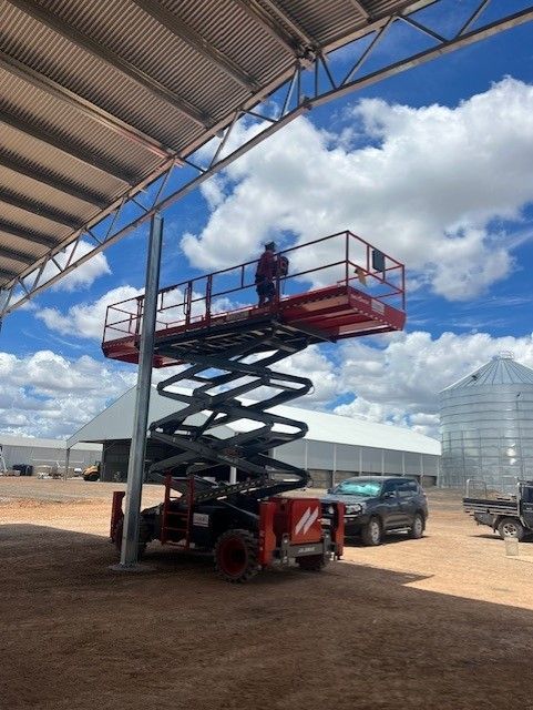 A person on a red scissor lift working on a metal post outdoors under a roof, near a farm building and parked vehicles.