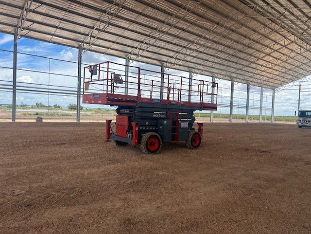 A red and grey scissor lift parked on a dirt floor inside a large, open-sided metal warehouse structure.