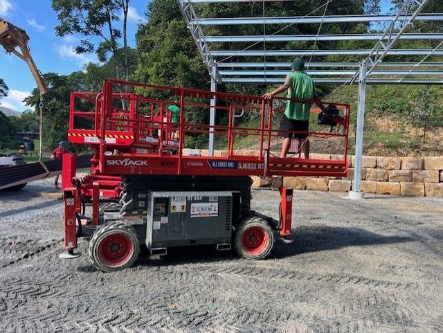 A construction worker stands on a red Skyjack scissor lift at an outdoor site with steel framework and a stone wall.