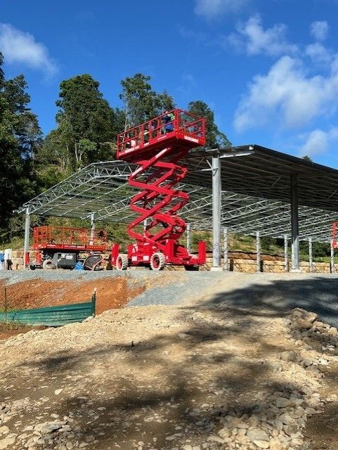 A red scissor lift is elevated under the metal frame of a large, unfinished building on a construction site.