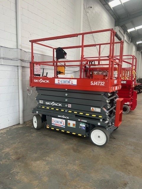 A red Skyjack SJ4732 electric scissor lift parked indoors on a concrete floor next to another red lift.