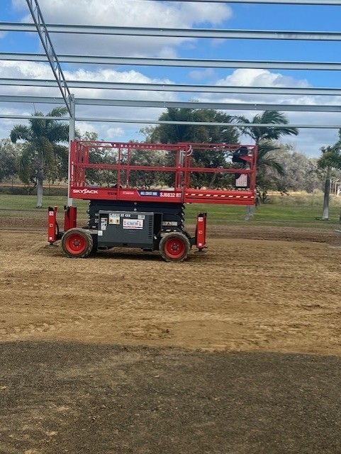 A red scissor lift parked on a dirt construction site under a partly cloudy sky with metal beams overhead.