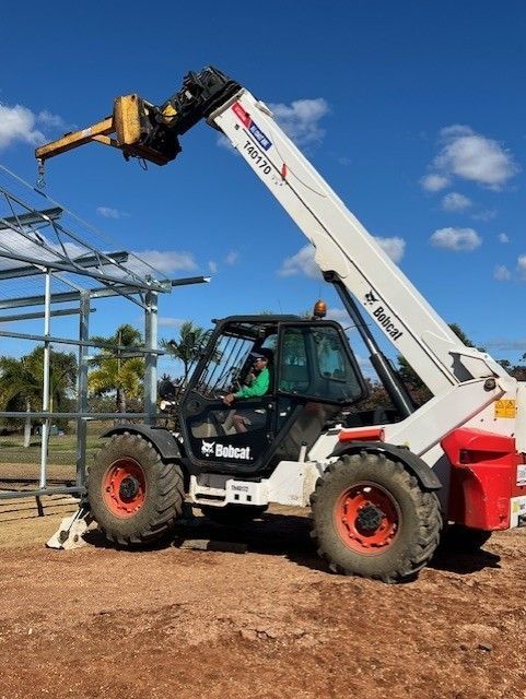 A Bobcat telehandler with an extended boom and crane attachment operates at a construction site under a blue sky.
