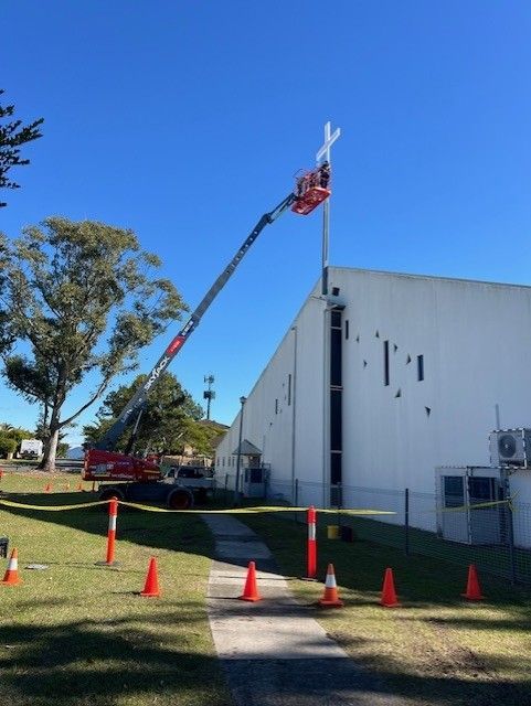 A person in a red cherry picker lift works on a white cross mounted to the roof of a building under a clear blue sky.