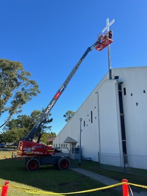 A person in a red boom lift working on a white cross atop the roof of a white building against a clear blue sky.