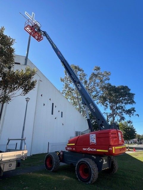 A red boom lift extends towards the top of a white building, with a person in the bucket working on a cross.