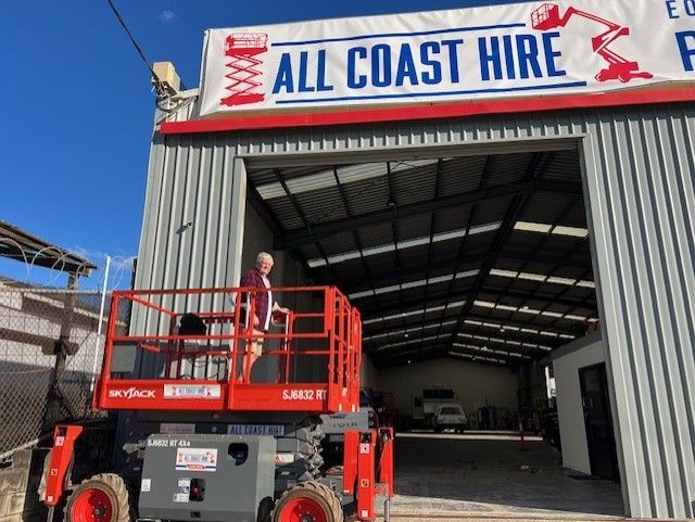 A person stands on a red scissor lift platform parked in front of a warehouse with an All Coast Hire sign above the door.
