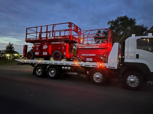 A white flatbed truck parked at dusk carrying two red scissor lifts on its bed.