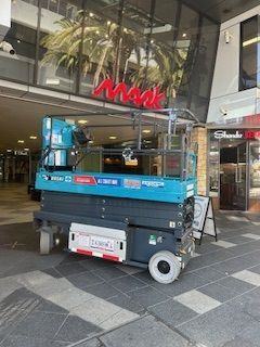A teal scissor lift parked on a tiled walkway in front of a building entrance with a red 
