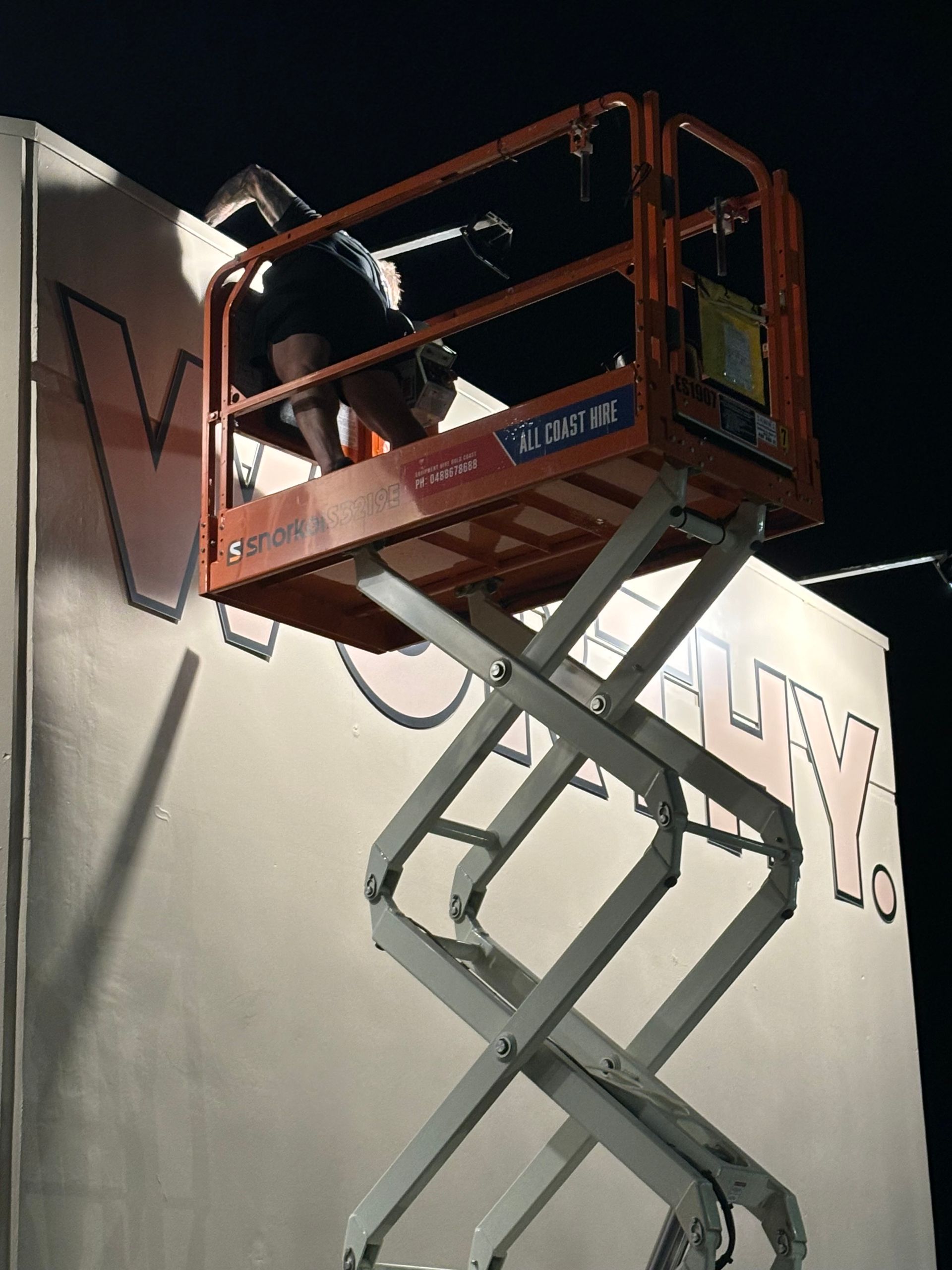 A person stands in a scissor lift platform at night, reaching toward the upper section of a building with painted letters.