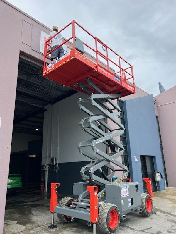 A person in a red and grey scissor lift working on a building exterior near an open warehouse bay.