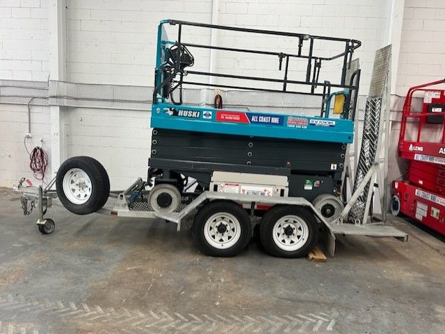 A teal scissor lift sits on a silver flatbed trailer parked inside a warehouse with a red scissor lift to the right.