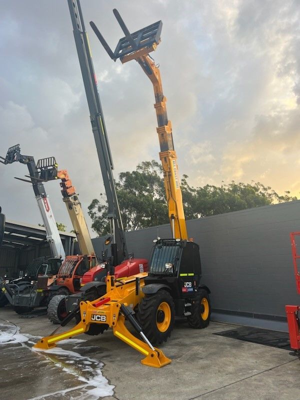 A yellow JCB telescopic handler with extended stabilizers and a raised boom parked on a concrete lot at dusk.