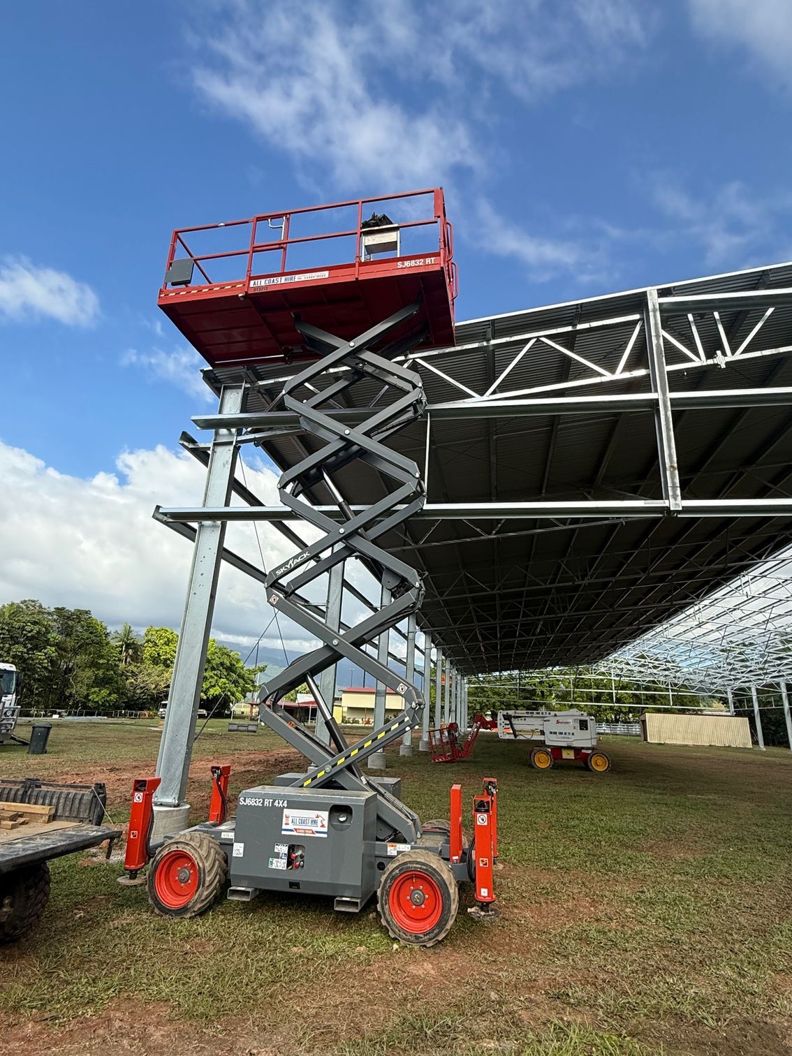 A red scissor lift is extended next to the metal frame of a large outdoor structure on a grassy lot under a blue sky.