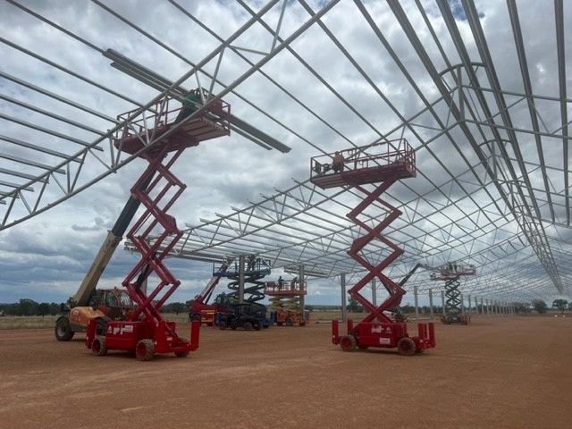 Workers in red scissor lifts install metal roof trusses on a large construction site under a cloudy sky.