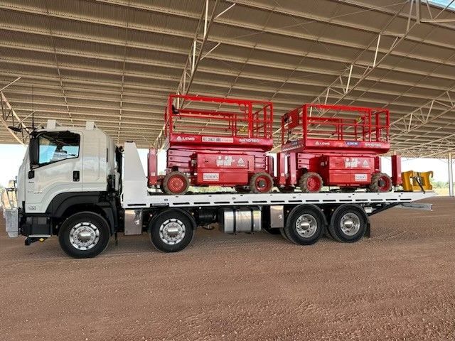 A white flatbed truck parked under a covered structure, carrying two red scissor lifts on its back.