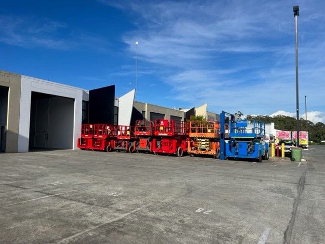 A row of red, orange, and blue scissor lifts parked in a concrete lot outside a warehouse under a clear blue sky.