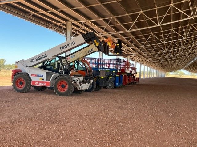 A Bobcat telehandler and several pieces of machinery parked under a large metal canopy on a gravel lot.