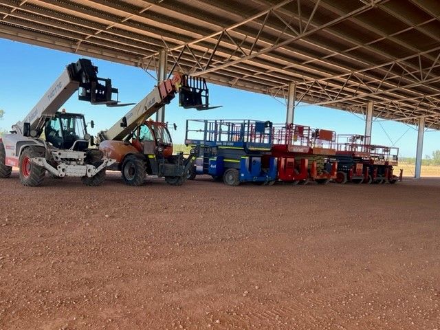 A row of construction equipment, including telehandlers and scissor lifts, parked under an industrial metal canopy.