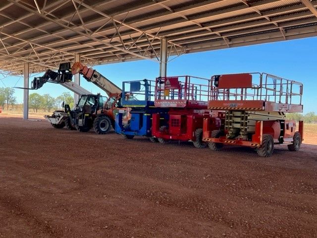 A telehandler and three scissor lifts parked in a row under a metal structure on a dirt lot.