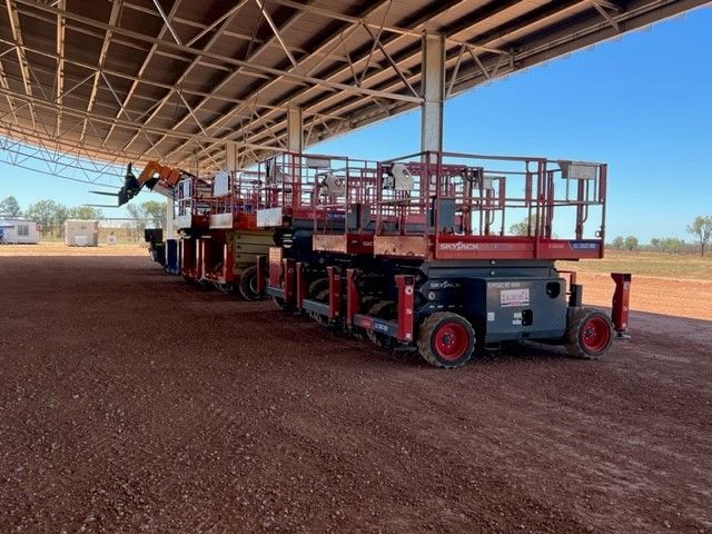 Several red scissor lifts parked under a metal canopy on a dirt lot under a clear blue sky.