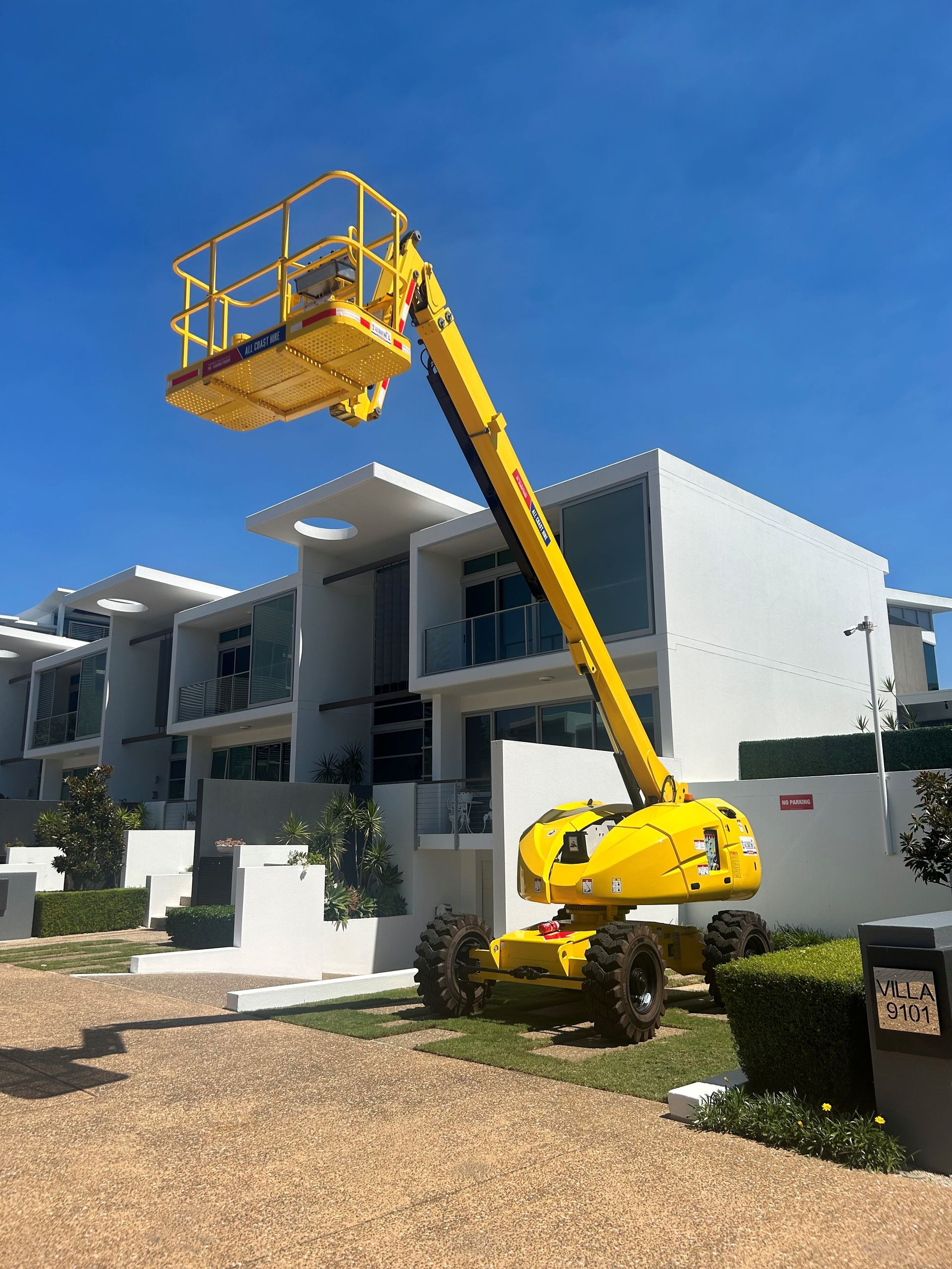 A yellow boom lift parked on a gravel area in front of modern white two-story townhouses under a clear blue sky.