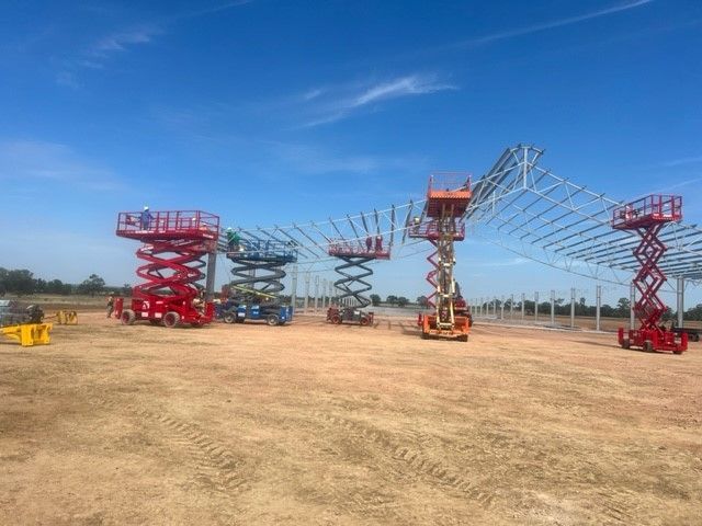 Five colorful scissor lifts arranged in a row on a dirt lot, supporting the metal framework of an unfinished structure.