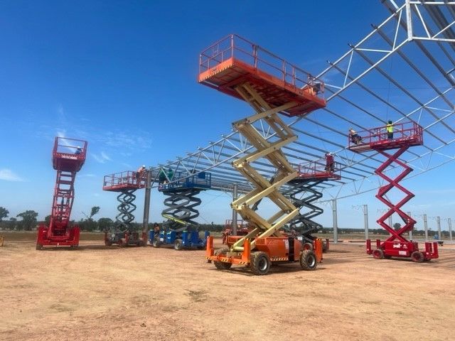 Multiple scissor lifts of varying colors work beneath a metal frame structure on a construction site under a clear sky.