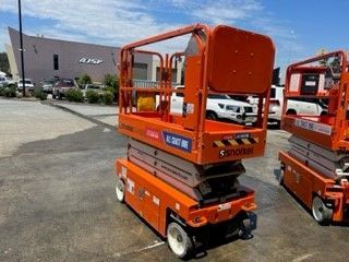 Two orange Snorkel scissor lifts parked in a lot outside a building on a sunny day.